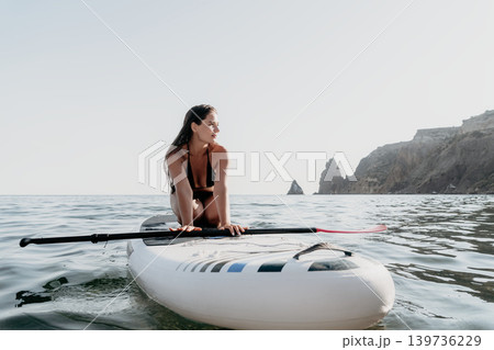 Woman Paddleboarding Ocean Coastline - A woman in a swimsuit kneels on a paddleboard in the ocean, looking towards a rocky coastline in the distance. Woman Paddleboarding Ocean Coastline - A woman in a swimsuit kneels on a paddleboard in the ocean, looking towards a rocky coastline in the distance. 139736229