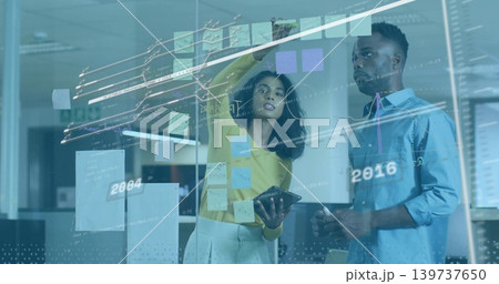 Office pair in yellow and blue arranging sticky notes on glass partition, holding tablet and marker Office pair in yellow and blue arranging sticky notes on glass partition, holding tablet and marker 139737650