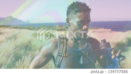 Standing hiker resting at coastal dune, holding blue water bottle, wearing dark tank top backpack Standing hiker resting at coastal dune, holding blue water bottle, wearing dark tank top backpack 139737888