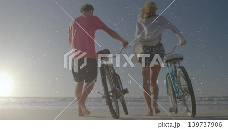 Walking couple wearing cover-up and pink tee pushing two bicycles on wet sand, low sun reflecting Walking couple wearing cover-up and pink tee pushing two bicycles on wet sand, low sun reflecting 139737906