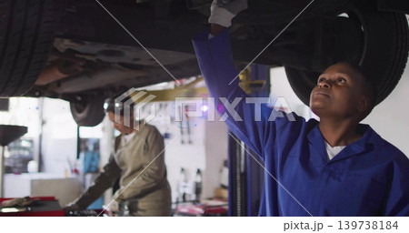 Inspecting technician in blue coveralls reaching under raised car at shop bay, tools, copy space Inspecting technician in blue coveralls reaching under raised car at shop bay, tools, copy space 139738184