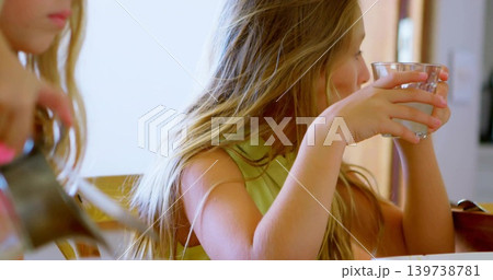 Holding glass, right girl wearing sleeveless top sipping at kitchen table with spoon, copy space Holding glass, right girl wearing sleeveless top sipping at kitchen table with spoon, copy space 139738781