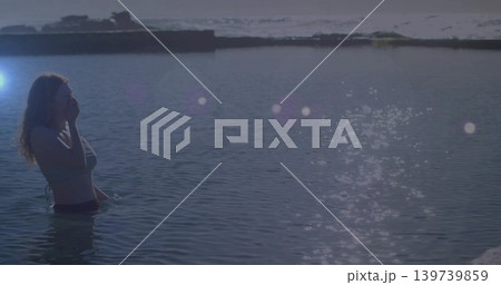 Standing woman wearing swimsuit touching face in shallow water near jetty, lens flare, copy space 139739859