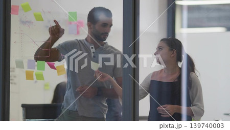 Writing man in grey polo and woman in dark dress charting on conference glass, sticky notes 139740003
