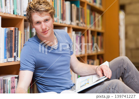 Adult male student sitting on library floor leaning against bookshelf and reading open textbook 139741227