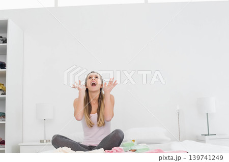 Woman sitting on bed in bright bedroom, wearing pink tank and gray leggings, raising hands Woman sitting on bed in bright bedroom, wearing pink tank and gray leggings, raising hands 139741249