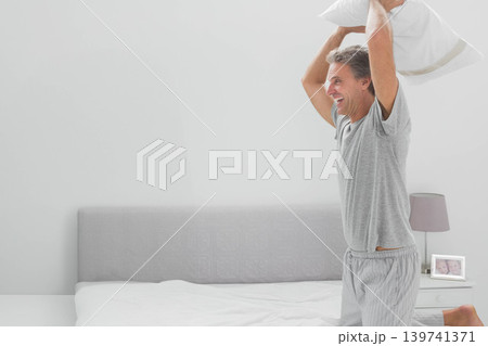 Man wearing gray T-shirt and striped pajama pants holding white pillow overhead on bed, copy space Man wearing gray T-shirt and striped pajama pants holding white pillow overhead on bed, copy space 139741371