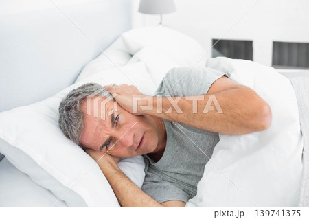 Mature male lying on side in bed wearing grey T-shirt holding ears near bedside lamp Mature male lying on side in bed wearing grey T-shirt holding ears near bedside lamp 139741375
