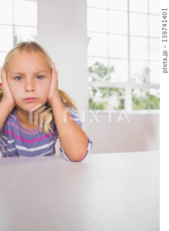Female child sitting at table pressing hands to ears in striped shirt, grid window, copy space Female child sitting at table pressing hands to ears in striped shirt, grid window, copy space 139741401