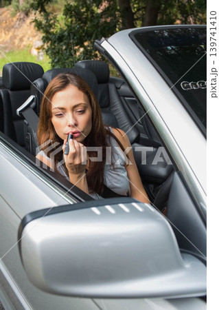 Middle-aged woman applying lipstick holding tube leaning toward side mirror in silver convertible Middle-aged woman applying lipstick holding tube leaning toward side mirror in silver convertible 139741410