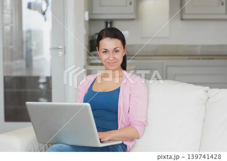 Woman sitting on white sofa holding silver laptop facing camera in kitchen with coffee maker Woman sitting on white sofa holding silver laptop facing camera in kitchen with coffee maker 139741428