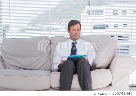 Man sitting on sofa in office waiting area holding blue folder, wearing white shirt, dark tie 139741446