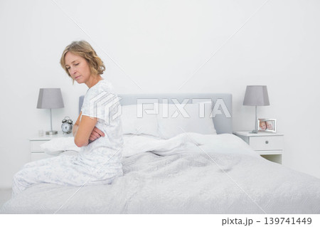Mature adult female sitting on bed edge, wearing patterned pajamas, beside alarm clock, copy space Mature adult female sitting on bed edge, wearing patterned pajamas, beside alarm clock, copy space 139741449