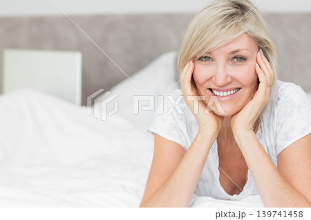 Mature woman reclining on bed, smiling, wearing pajama top, showing ring, bedside book, copy space Mature woman reclining on bed, smiling, wearing pajama top, showing ring, bedside book, copy space 139741458