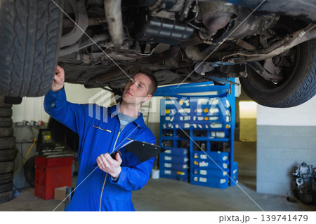 Male mechanic inspecting car undercarriage on hydraulic lift while holding clipboard in shop Male mechanic inspecting car undercarriage on hydraulic lift while holding clipboard in shop 139741479