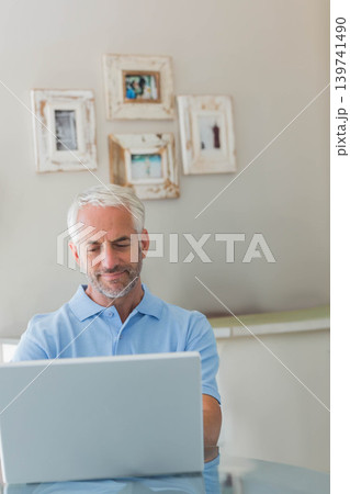 Senior man in blue polo shirt sitting at glass table at home, working on silver laptop Senior man in blue polo shirt sitting at glass table at home, working on silver laptop 139741490