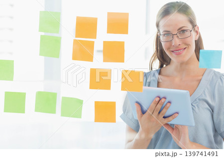 Mid-adult female holding tablet by glass with orange green sticky notes in office wearing glasses 139741491