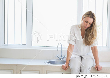 Mid-adult female sitting on countertop near sink, wearing white top, hands resting, copy space Mid-adult female sitting on countertop near sink, wearing white top, hands resting, copy space 139741524
