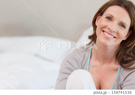 Middle-aged woman sitting on bed, smiling at camera, wearing cardigan and pale blue camisole Middle-aged woman sitting on bed, smiling at camera, wearing cardigan and pale blue camisole 139741538