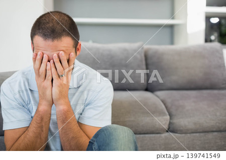 Mid-adult man sitting on gray sofa in living room covering face, wedding band visible, copy space Mid-adult man sitting on gray sofa in living room covering face, wedding band visible, copy space 139741549