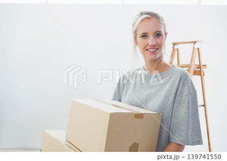 Woman in her 20s wearing grey T-shirt holding taped moving boxes by wooden ladder, copy space 139741550
