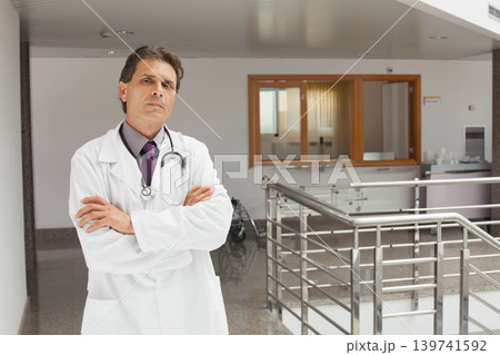 Midlife male doctor standing with crossed arms in hospital in lab coat with stethoscope, copy space Midlife male doctor standing with crossed arms in hospital in lab coat with stethoscope, copy space 139741592