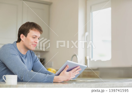 Man sitting in kitchen at stone countertop holding tablet with coffee mug and bananas, copy space Man sitting in kitchen at stone countertop holding tablet with coffee mug and bananas, copy space 139741595