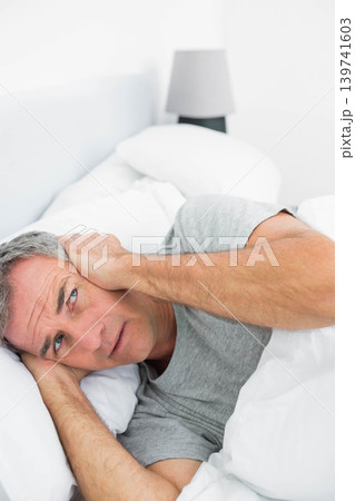 Mature man lying in bedroom on bed with pillow pressing hand to ear, wearing gray T-shirt Mature man lying in bedroom on bed with pillow pressing hand to ear, wearing gray T-shirt 139741603