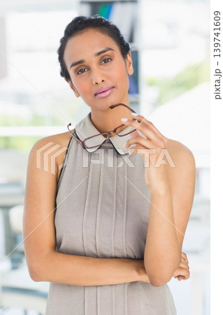 Asian woman posing while holding tortoise glasses near face in sleeveless collared dress at office Asian woman posing while holding tortoise glasses near face in sleeveless collared dress at office 139741609