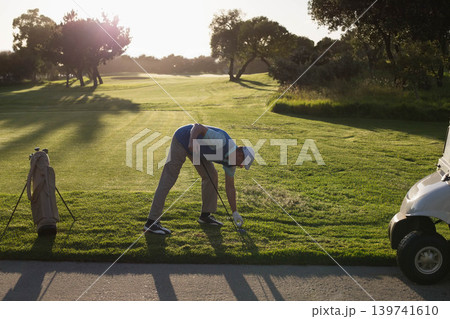 Mature man bending, placing golf ball on tee at sunlit course with white cap glove bag Mature man bending, placing golf ball on tee at sunlit course with white cap glove bag 139741610