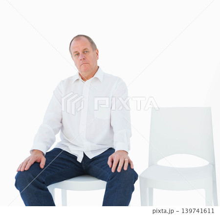 Senior man sitting on white chair in studio, empty chair at right, wearing white shirt, jeans Senior man sitting on white chair in studio, empty chair at right, wearing white shirt, jeans 139741611