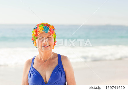 Senior woman standing, posing on sandy shore in blue swimsuit, floral cap, ocean waves horizon Senior woman standing, posing on sandy shore in blue swimsuit, floral cap, ocean waves horizon 139741620