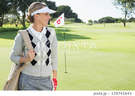 Female golfer standing on green by flagstick and hole carrying bag while wearing visor, copy space Female golfer standing on green by flagstick and hole carrying bag while wearing visor, copy space 139741643