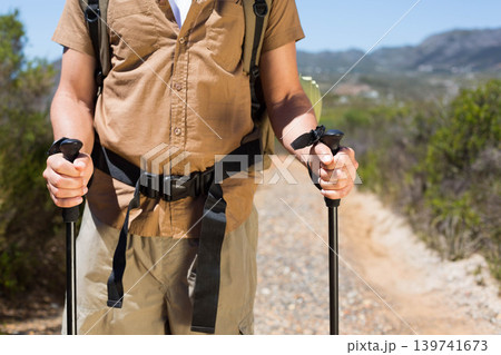 Male hiker standing on trail holding black trekking poles, wearing brown shirt and backpack Male hiker standing on trail holding black trekking poles, wearing brown shirt and backpack 139741673