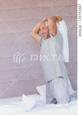 Senior male kneeling on bed pulling white shirt over head wearing grey T-shirt and striped pajamas Senior male kneeling on bed pulling white shirt over head wearing grey T-shirt and striped pajamas 139741687