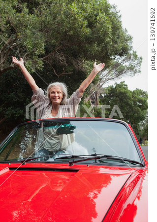 Senior woman standing through red convertible raising arms smiling in shawl and jeans on park road 139741692