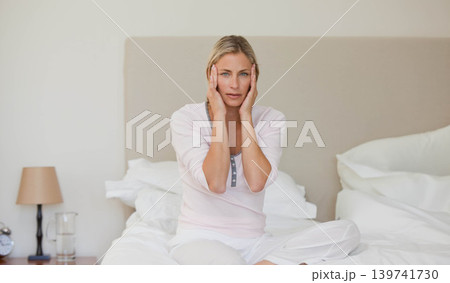 30s woman in pajamas sitting on bed holding cheeks, bedside table with lamp and alarm clock 30s woman in pajamas sitting on bed holding cheeks, bedside table with lamp and alarm clock 139741730
