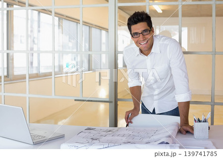 Man in white shirt leaning over desk in office studying plans with pen, laptop, copy space 139741785