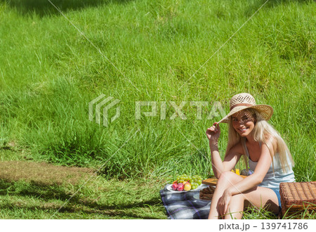 Woman sitting on checked blanket grassy slope holding straw hat and fruit basket beside, copy space 139741786