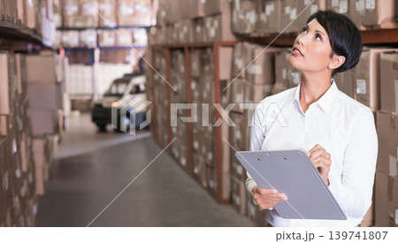 Mature woman in white shirt holding clipboard, pen checking boxes in storage aisle, copy space Mature woman in white shirt holding clipboard, pen checking boxes in storage aisle, copy space 139741807