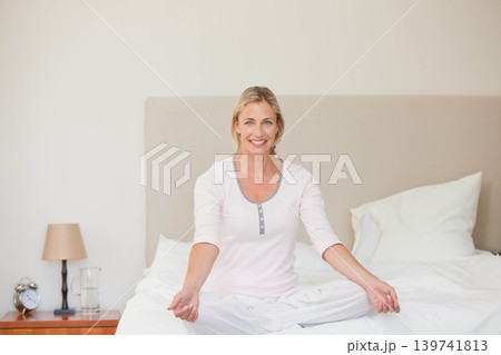 Middle-aged woman sitting cross-legged on bed at home wearing pale pink pajamas near nightstand Middle-aged woman sitting cross-legged on bed at home wearing pale pink pajamas near nightstand 139741813