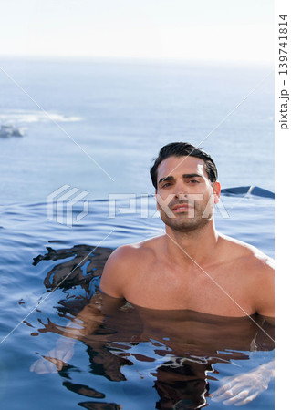 Man standing chest-deep in infinity pool, shirtless, facing lens with ocean horizon, rock left 139741814