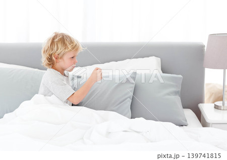 Child boy sitting on bed holding small object in right hand amid white duvet, copy space 139741815