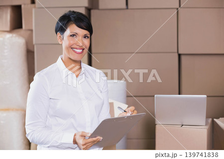 Mature woman standing in storage area wearing white shirt holding clipboard and pen near laptop Mature woman standing in storage area wearing white shirt holding clipboard and pen near laptop 139741838