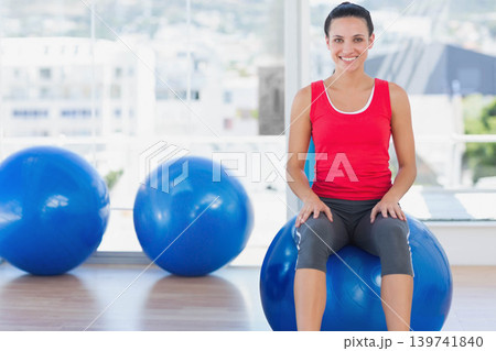 Female sitting on blue exercise ball smiling in studio with large windows, copy space Female sitting on blue exercise ball smiling in studio with large windows, copy space 139741840