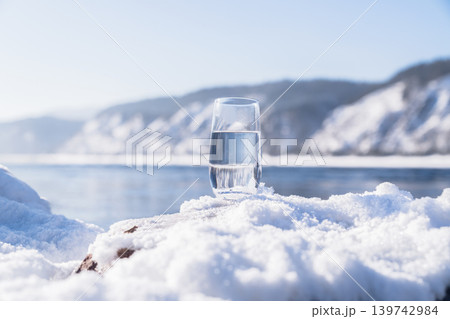 A glass filled with water stands on a snow-covered surface near a river during winter. Mountains rise in the background under a clear blue sky. A clean glass of healthy mineral water stands in the A glass filled with water stands on a snow-covered surface near a river during winter. Mountains rise in the background under a clear blue sky. A clean glass of healthy mineral water stands in the 139742984