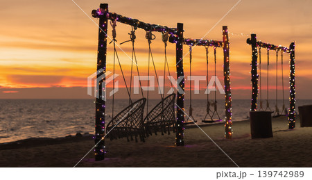 Swings on a beach are illuminated by lights as the sun sets over the sea. The sky shows warm colors while waves gently touch the shore. High quality photo 139742989