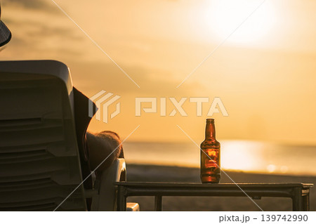 A bottle of beer on the table during sunset at sea. A man looks at the sea, rests and watches the setting sun, the concept of a holiday at a resort. Relaxing and enjoying a drink in the evening. High 139742990