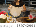 A woman prepares sweet roti at a Bangkok street stall, skillfully stretching the dough. She has gloves on her hands while oils and other ingredients are nearby. High quality photo 139742991