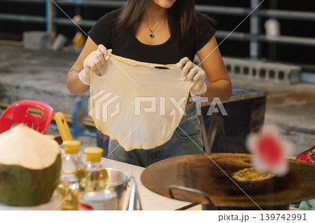 A woman prepares sweet roti at a Bangkok street stall, skillfully stretching the dough. She has gloves on her hands while oils and other ingredients are nearby. High quality photo 139742991
